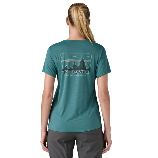 En kvinde med tilbagestrøget blondt hår er iført en Patagonia W's Cap Cool Daily Graphic Shirt, Dame, 73 Skyline/Wetland Blue X-Dye-hurtingtørrende med UPF-beskyttelse - og grå bukser - og ser væk fra kameraet på en hvid baggrund.