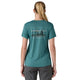 En kvinde med tilbagestrøget blondt hår er iført en Patagonia W's Cap Cool Daily Graphic Shirt, Dame, 73 Skyline/Wetland Blue X-Dye-hurtingtørrende med UPF-beskyttelse - og grå bukser - og ser væk fra kameraet på en hvid baggrund.