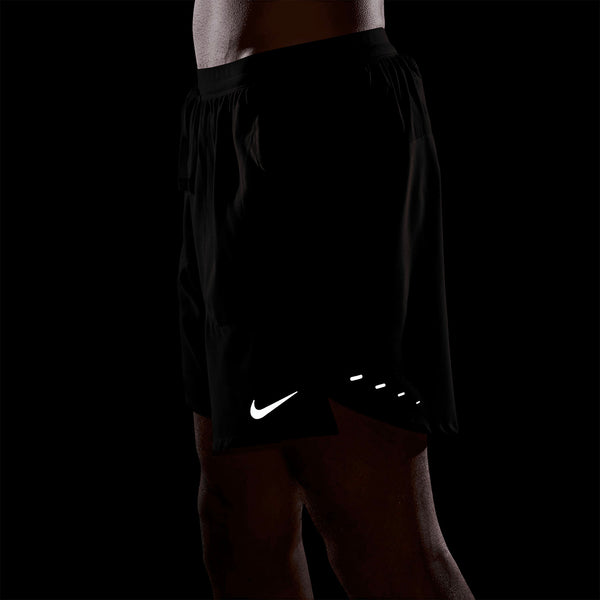 En person ses fra taljen til midt på låret iført Nike Stride 7" Brief-Lined Running Shorts til mænd i Black/Reflective Silver med det ikoniske Nike-logo nederst på venstre ben mod en sort baggrund.