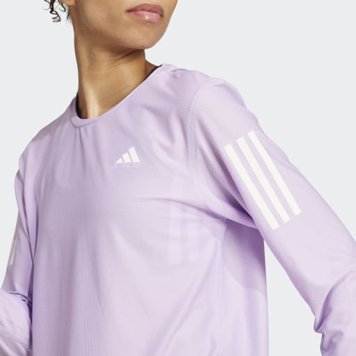 En kvinde er iført Adidas Otr B Ls langærmet top i lyslilla (Powplu) med hvide ærmestriber, Adidas-logo på brystet og AEROREADY-teknologi. Hendes ansigt er delvist synligt mod en almindelig hvid baggrund.