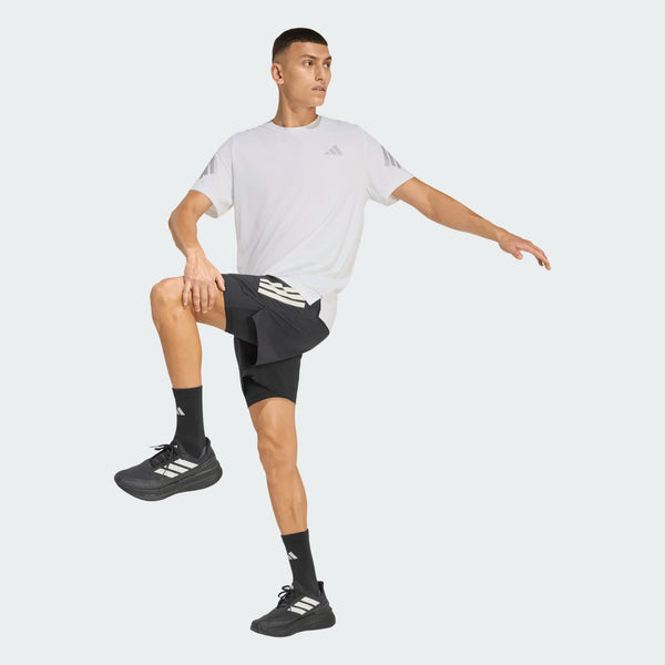En mand i Adidas Adi365 Running Tee, Herre, White, sorte shorts og sorte sportssko står med det ene knæ løftet og den ene arm strakt i en opvarmningspositur mod en almindelig lys baggrund.
