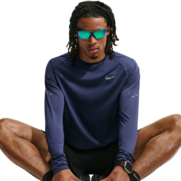 En person med langt hår, iført en Nike Dri-FIT ADV langærmet løbetrøje til mænd i farven Midnight Navy/Reflective Silver, sorte shorts, reflekterende solbriller og et smartwatch, sidder med benene krydsede foran en hvid baggrund.