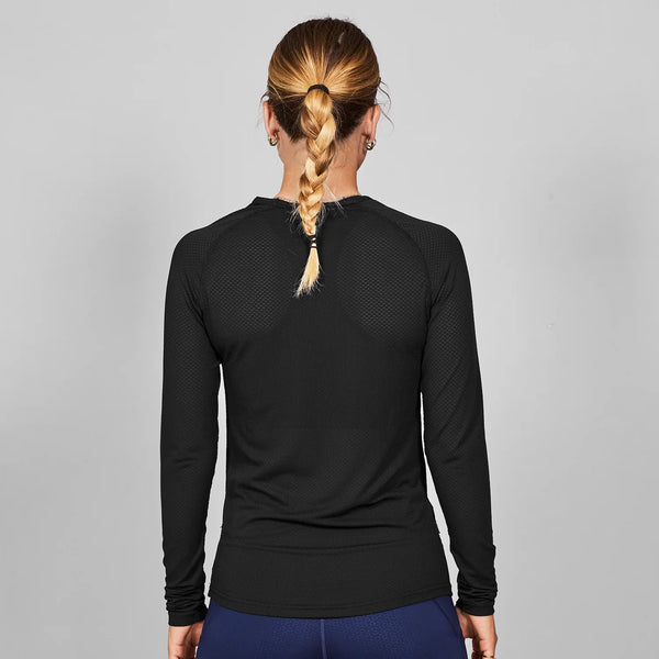 En person med en enkelt fletning kigger væk fra kameraet og er iført Saysky Mesh Base Layer Long Sleeve i sort (unisex) sammen med blå bukser på en almindelig grå baggrund.