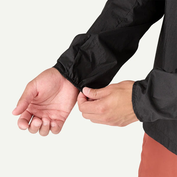 En person justerer den elastiske manchet på en Clement Blue Patagonia Houdini Jkt, Herre, med sin venstre hånd. De er iført rustfarvede bukser mod en almindelig råhvid baggrund.