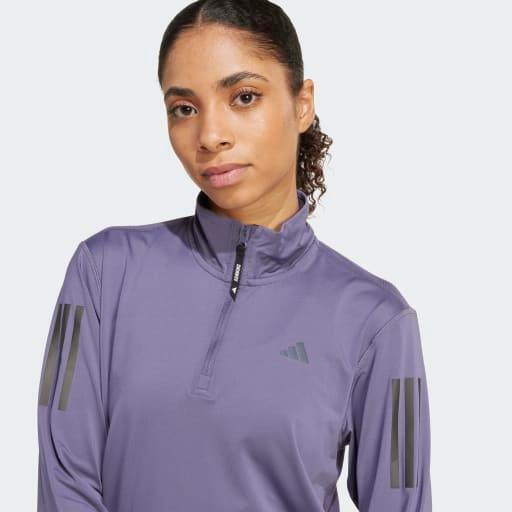 En kvinde med tilbagestrøget krøllet hår bærer Adidas Otr B Hzip træningstrøje for damer i lilla med sorte ærmestriber, et lille logo og AEROREADY-materiale. Previos-toppen med halv lynlås vises, mens hun kigger fremad mod en ensfarvet baggrund.