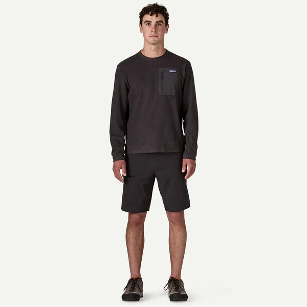 En mand står med front mod en hvid baggrund iført en sort Patagonia R1 Air Crew-pullover til mænd lavet af genbrugspolyester, sammen med sorte knælange shorts og sorte sneakers.