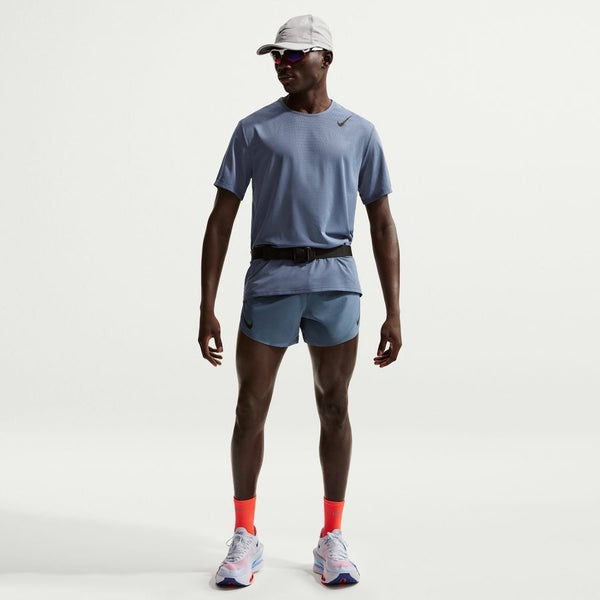 En mand ser frem for sig i grå kasket, solbriller, Nike Dfadv Aroswft SS Top til mænd i Diffused Blue/Black, matchende shorts, bæltetaske, orange sokker og hvide løbesko på en almindelig hvid baggrund.