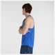 En person med kort brunt hår står sidelæns med armene over kors iført en New Balance Athletics Tank, Herre, Blue Gemstone Heather. Navy shorts og en lille tatovering på venstre overarm fuldender looket mod en almindelig hvid baggrund.