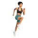 En kvinde i sort sports-bh, Adidas OTR Short Tights 6" Dame PRETEA, hvide sokker og hvide sneakers er fanget midt i et spring med et løftet knæ og bøjede arme mod en almindelig hvid baggrund.