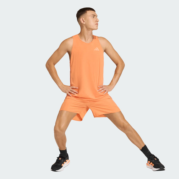 En person i orange Adidas Adi365 Singlet, Herre, Duor og matchende shorts står sidelæns med spredte ben og hænderne på hofterne. De bærer sorte sokker og sorte sportssko med orange detaljer. Baggrunden er almindelig lysegrå.