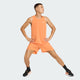 En person i orange Adidas Adi365 Singlet, Herre, Duor og matchende shorts står sidelæns med spredte ben og hænderne på hofterne. De bærer sorte sokker og sorte sportssko med orange detaljer. Baggrunden er almindelig lysegrå.