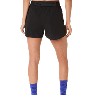 Person vist fra taljen og ned, iført Asics Metarun Split Shorts til kvinder i Performance Black, parret med blåmønstrede strømper, stående mod en almindelig hvid baggrund.