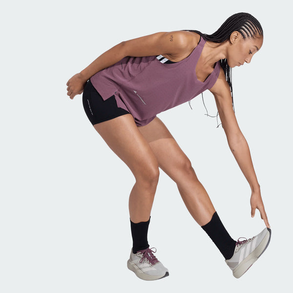 En person iført Adidas Adi365 Hk Singlet til kvinder, sorte shorts, sorte sokker og grå sportssko læner sig frem og rører ved sin venstre tå med højre hånd, mens han strækker sig mod en almindelig lys baggrund.