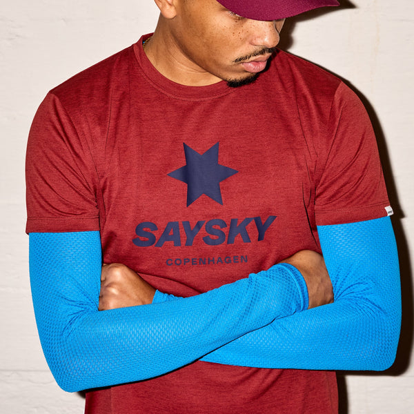 En person er iført en rød Saysky Logo Combat Pro T-shirt og en matchende kasket med en marineblå stjerne, lyseblå ærmer indenunder og har armene over kors.