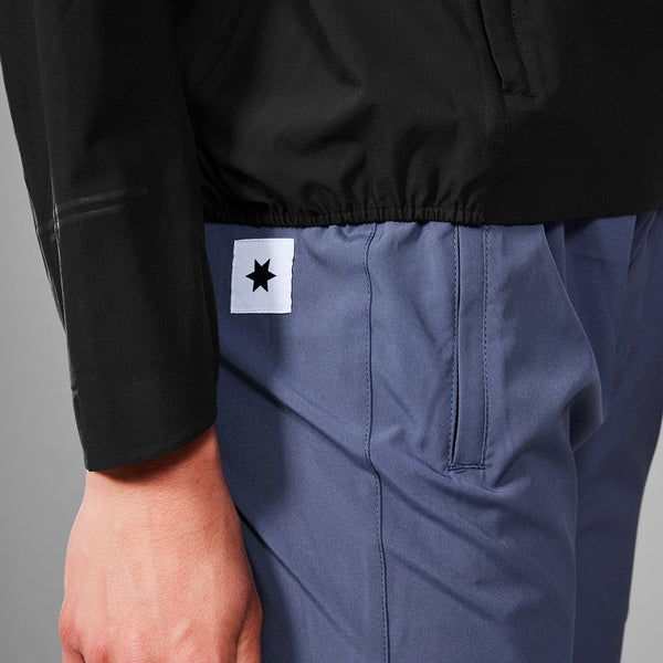 En person er iført blå bukser med et hvidt rektangulært mærke med en sort stjerne ved lommen og en Saysky Clean Pace Flex Hooded Jacket, Dame, Black. Kun underkroppen og den ene arm er synlig.