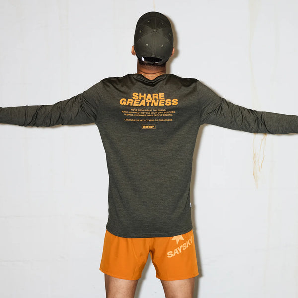 En person står med udstrakte arme iført en grøn Saysky Statement Pace Long Sleeve til mænd med "SHARE GREATNESS" i orange, parret med knaldorange shorts for et behageligt og modigt look.