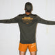 En person står med udstrakte arme iført en grøn Saysky Statement Pace Long Sleeve til mænd med "SHARE GREATNESS" i orange, parret med knaldorange shorts for et behageligt og modigt look.