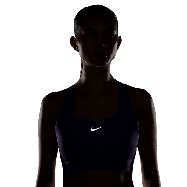 En kvinde iført en lilla Nike High-Support Lightly Lined Sports Bra (Dame, Purple Pulse) med et reflekterende logo står foran en hvid baggrund. Hendes ansigt og krop er stort set i skygge på grund af den bagfra kommende belysning, hvilket fremhæver bh’ens design med høj støtte – ideel til løbetræning eller andre aktiviteter med høj belastning.