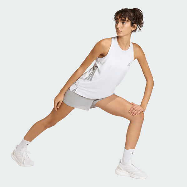 Iført Adidas Iconic Tank Top, Dame, White, lysegrå shorts, hvide sokker og adidas-sneakers springer en person sidelæns med et bøjet og et strakt ben mod en almindelig lys baggrund.