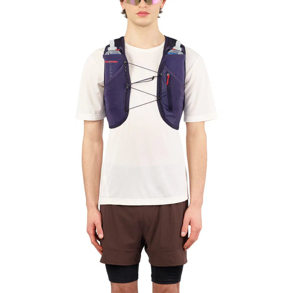 En person iført solbriller, en hvid kortærmet skjorte, brune shorts med sorte leggings indenunder og et Salomon Active Skin 4 Set (Unisex) i Maritime Blue/Astral Aura/Haute Red - ideelt til trailløb.