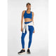 En kvinde kigger fremad iført en New Balance Define Train Bra, Dame, i Galaxy Blue med matchende leggings, sorte sneakers, en lysegrå sweatshirt bundet i taljen og en hvid baseballkasket på en almindelig hvid baggrund.
