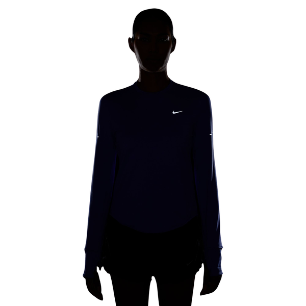 En person står med ansigtet vendt fremad, med silhuetten aftegnet mod en hvid baggrund. Personen har en mørk lilla Swift UV langærmet løbetrøje fra Nike på, med et reflekterende sølvfarvet logo på brystet, samt mørke shorts. Ansigtstrækkene er i skygge.