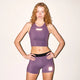 En kvinde med langt krøllet hår iført en ærmeløs crop top og Saysky W Flow+ Race Short Tights 4" i lilla står op ad en almindelig lys væg med hænderne på hofterne og kigger ind i kameraet.