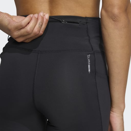 En kvinde kigger væk fra kameraet iført Adidas Adi365 Leggins Dame i sort med en lynlåslomme og "CLIMALWARM" på siden. Hendes venstre hånd hviler bag hendes ryg og fremhæver disse slanke leggins med høj talje.
