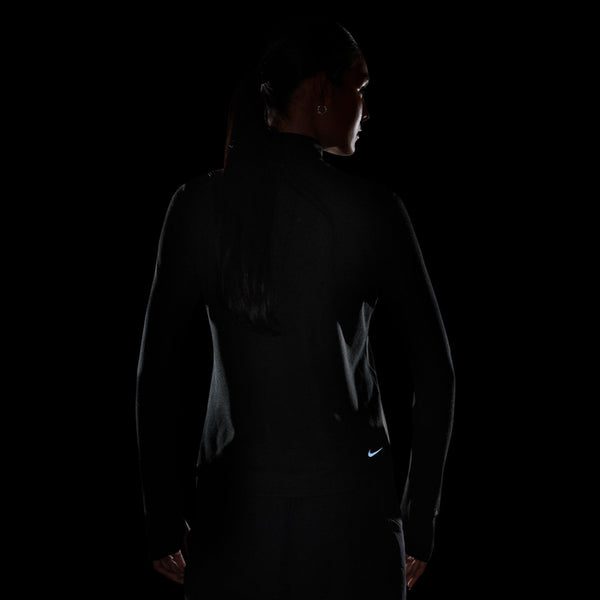 En person med langt hår, set bagfra i et svagt oplyst rum, er iført Nike ACG Half Zip Running Top til kvinder i Black/Black/Summit White. Nike-logoet ses nederst på ryggen, omkranset af diskrete highlights.