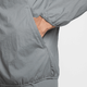 En hånd er delvist inde i lommen på en Nike Repel UV Running Jacket, Herre, Smoke Grey/Smoke Grey/Reflective Silv, set fra siden mod en ensfarvet baggrund.