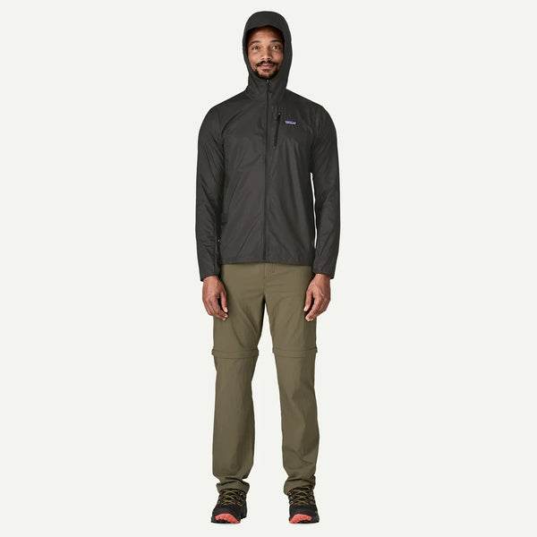 En person står og kigger fremad iført en Patagonia Houdini Jacket, Herre, Black w/Black, olivengrønne bukser og vandresko med orange detaljer på en almindelig hvid baggrund.