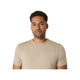 En mand med kort krøllet hår og et trimmet skæg bærer Asics Road Seamless Ss Top til mænd i creme, en kortærmet t-shirt, mens han kigger fremad mod en almindelig hvid baggrund.