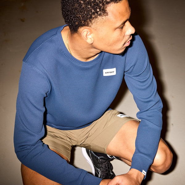En mand i en blå Saysky Motion Waffle Long Sleeve, Herre, lysebrune shorts og sorte sneakers knæler og kigger til højre. Den teknisk langærmet trøje har et lille hvidt brystmærke. Den ensfarvede baggrund passer til løbeentusiaster.