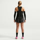 En person vender ansigtet væk fra kameraet og er iført en sort/hvid Nike AeroSwift Running Singlet (dame), sorte ærmer, en mørk plisseret nederdel, sorte knæstrømper og pink sportssko på en almindelig lys baggrund.
