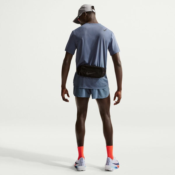 En person står med ryggen til og er iført en blå Nike Dfadv Aroswft Ss Top, Herre, Diffused Blue/Black og blå shorts, med en grå kasket, sort bæltetaske og hvide løbesko med røde sokker. Baggrunden er almindelig hvid.