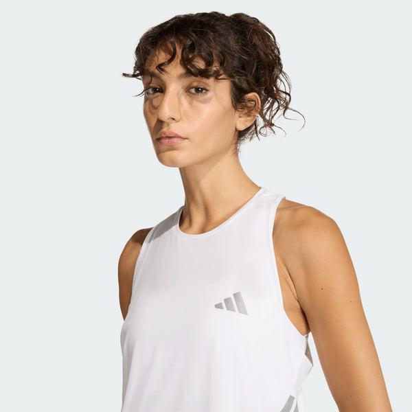 En kvinde med krøllet brunt hår bærer Adidas Iconic Tank Top, Dame, White - en ærmeløs sportstop med et gråt logo - mod en almindelig lysegrå baggrund, mens hun kigger lidt forbi kameraet.
