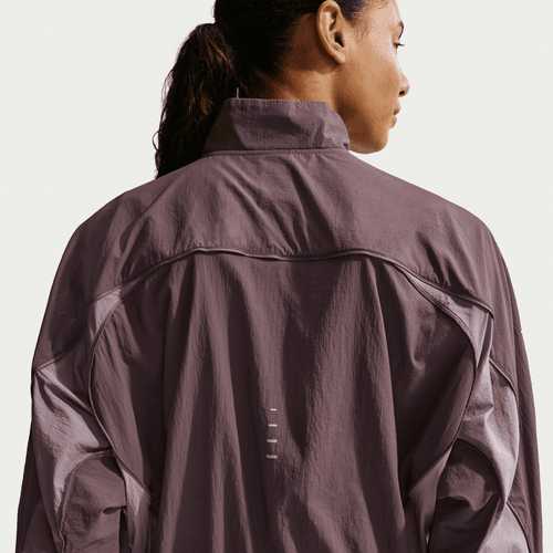 En person med langt, mørkt hår bundet tilbage ses bagfra iført Nike Swift Repel UV Protection Running Jacket til kvinder i Tattoo/LT Violet Ore/Reflective Silver med høj krave og lodrette refleksdetaljer på ryggen.