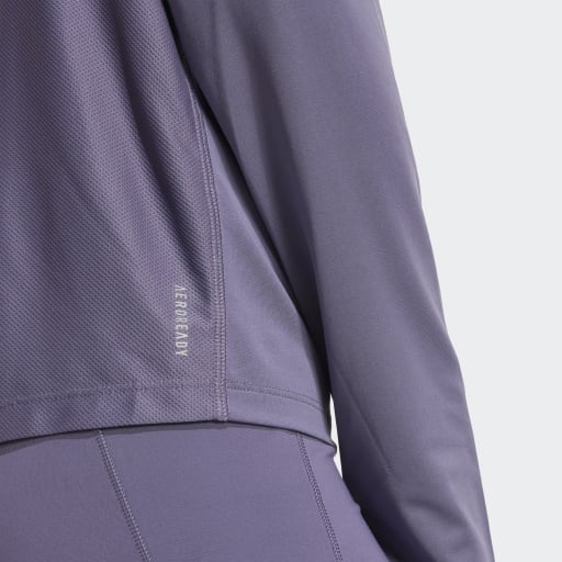 Et nærbillede viser en person i en matchende lavendelfarvet Adidas Otr B Hzip træningstrøje dame og leggings. Den langærmede top har "AEROREADY" trykt nær kanten, hvilket fremhæver det fugtabsorberende stof.