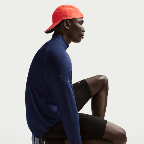 En person i en Nike Stride Half Zip Midlayer Running Top til mænd, Midnight Navy/Reflective Silver, sidder i sideprofil mod en almindelig lys baggrund, sammen med sorte sportsshorts og en knaldrød baglæns kasket.