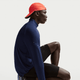 En person i en Nike Stride Half Zip Midlayer Running Top til mænd, Midnight Navy/Reflective Silver, sidder i sideprofil mod en almindelig lys baggrund, sammen med sorte sportsshorts og en knaldrød baglæns kasket.