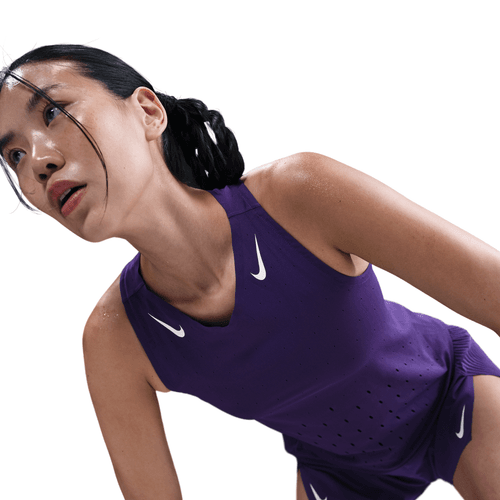 En kvinde i Nike AeroSwift Running Singlet, Dame, Court Purple/White læner sig frem og ser træt og svedig ud med hvide Nike-logoer synlige på en hvid baggrund.