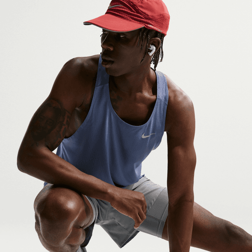 En person i rød kasket, grå shorts, trådløse øretelefoner og Nike Nk Df Fast Singlet til mænd i Diffused Blue/Reflective Silver poserer mod en hvid baggrund og fremhæver stilfuldt herre træningstøj.