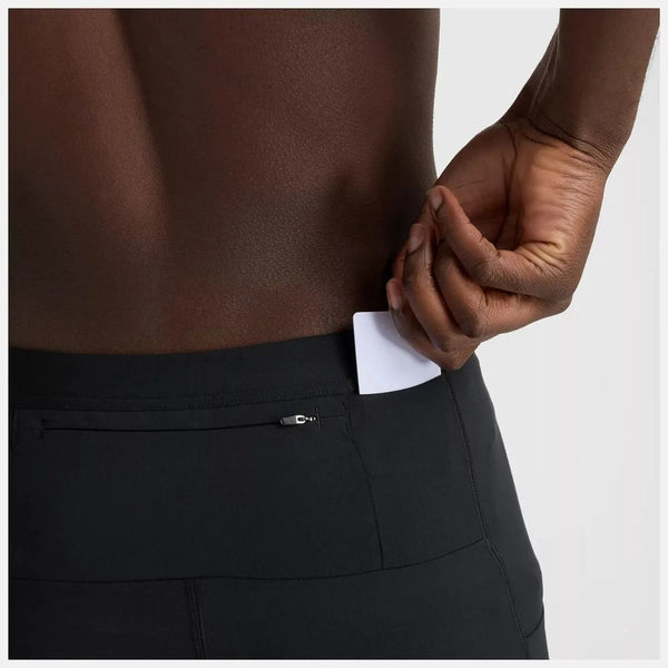 En mand stikker et hvidt kort ned i den lille lynlåslomme bag på den sorte NB Sleek Pocket Half Tight 9" fra New Balance. Det beskårne billede fremhæver kun hans nedre del af ryggen og hånden.