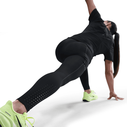 En person i Nike Therma-FIT ADV High-Waisted 7/8 Reflective Running Leggings (Dame, Black/Dk Smoke Grey) og neongrønne sneakers laver et gulvstræk med den ene arm oppe og den anden nede, set fra en lav vinkel bagfra, der fremhæver deres atletiske udstyr.