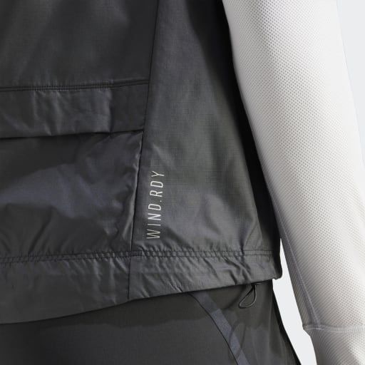 Nærbillede af Adidas Otr B Vest til kvinder i sort med WIND.RDY-tekst ved en søm, lagt over et hvidt tekstureret ærme og sort løbetøj til kvinder.