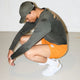 En person bærer en Saysky Statement Pace Long Sleeve, Herre, Green skjorte med orange shorts, hvide sokker og hvide sneakers, mens han sidder på hug på et lyst gulv mod en almindelig hvid væg. Skjorten sikrer svedtransporterende komfort til aktive.