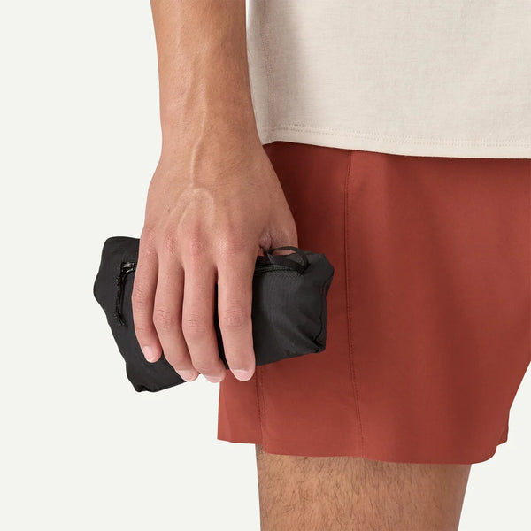 En person i rustne shorts og en beige skjorte holder en lille sort pose med lynlås - ideel til at pakke Patagonia Houdini Jkt, Herre, Clement Blue - i sin højre hånd.