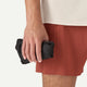En person i rustne shorts og en beige skjorte holder en lille sort pose med lynlås - ideel til at pakke Patagonia Houdini Jkt, Herre, Clement Blue - i sin højre hånd.