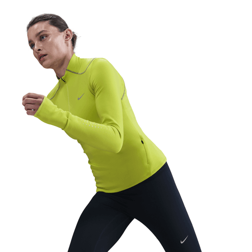 En kvinde iført en lys Cyber Therma-FIT ADV 1/2-Zip Reflective Running Top fra Nike og sorte leggings ses i gang mod en hvid baggrund og ser ud til at løbe eller træne.