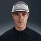 En person iført Nike Unstructured Swoosh Cap i Medium Ash/College Grey/Sail sammen med en mørk jakke med lynlås kigger ind i kameraet med et neutralt udtryk mod en mørk baggrund med gradient.